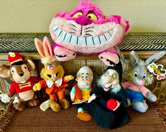 May include: A collection of five plush toys including a large pink Cheshire Cat, a mouse in a red uniform, a rabbit in an orange suit, a small elderly character in a vest, a witch in a black robe holding a red apple, and a grey rabbit in a pink shirt.