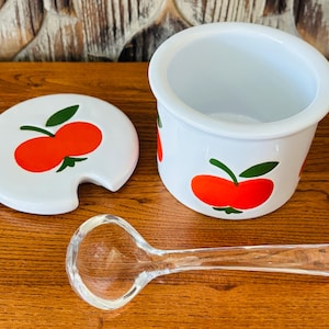 May include: A white ceramic jar with a lid and a clear glass spoon. The jar and lid are decorated with red apple designs with green leaves. The spoon is clear and has a rounded bowl.