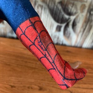 K&ouml;nnte beinhalten: Nahaufnahme eines blauen und roten Spiderman-Kost&uuml;mbeins. Der rote Teil hat ein schwarzes Netzdesign. Das Bein liegt auf einer Holzoberfl&auml;che.
