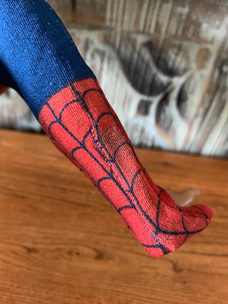 K&ouml;nnte beinhalten: Nahaufnahme eines blauen und roten Spiderman-Kost&uuml;mbeins. Der rote Teil hat ein schwarzes Netzdesign. Das Bein liegt auf einer Holzoberfl&auml;che.