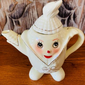 May include: A white ceramic teapot shaped like a person with a smiling face, wearing a hat and a bow tie. The teapot has a gold trim around the hat and bow tie.