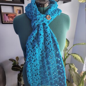 May include: A turquoise crochet scarf with a wooden button closure. The scarf is displayed on a mannequin and features a textured, openwork design. The scarf is a vibrant shade of blue and appears handmade.