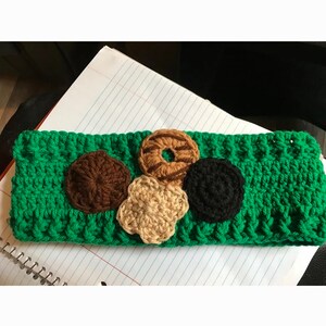 May include: A bright green crocheted headband featuring four circular appliqués in shades of dark brown, light brown, tan, and black. The headband has a textured, ribbed stitch pattern and is displayed horizontally against a white lined paper background.