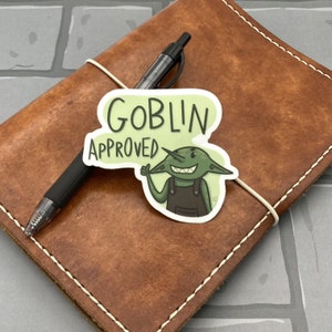 May include: A brown leather journal with a white elastic closure, a black pen, and a sticker that says "GOBLIN APPROVED." The sticker features a green goblin giving a thumbs up.
