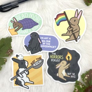 May include: Set of nine stickers featuring various illustrations of rabbits and bunnies. The stickers have different backgrounds and text, including "Read More Banned Books", "Eat the Rich", "The Horrors Persist", and "I'm not a big fan of the government".