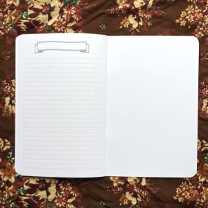 May include: Open notebook with lined pages and a blank right page. A banner illustration is at the top of the left page. The notebook is open on a floral patterned brown surface.