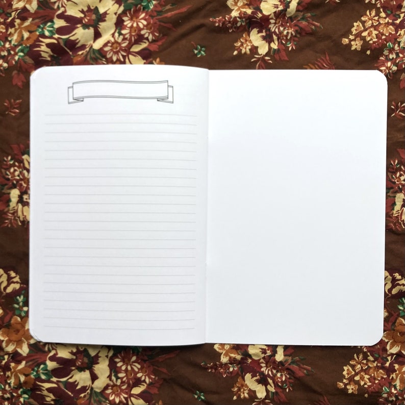 May include: Open notebook with lined pages and a blank right page. A banner illustration is at the top of the left page. The notebook is open on a floral patterned brown surface.