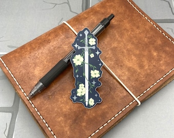 May include: A sticker featuring a silver sword with a white flower vine wrapped around it. The sword is on a dark blue background with white stars. The sticker is on a brown leather journal.