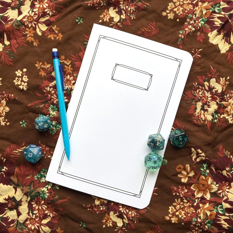 May include: A blank white notebook with a black border and a rectangular label area, next to a blue pencil and several teal and glittery polyhedral dice. The items are arranged on a brown floral patterned fabric.