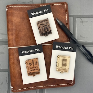 May include: Three wooden pins featuring illustrations of book covers. The pins are labeled "Dungeon Master's Guide", "Player's Handbook", and "Monster Manual".