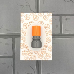 May include: A gray and orange ink stamp with a 20-sided die design. The stamp is surrounded by a white background with a pattern of 20-sided dice.