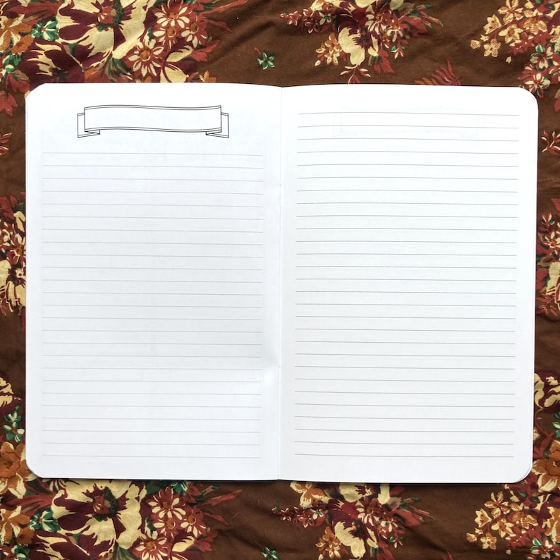 May include: An open, lined notebook with a banner graphic at the top of the left page. The notebook is white, and the lines are gray. The background is a brown fabric with a floral pattern.