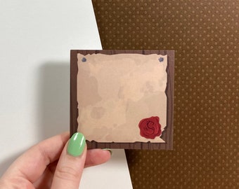 May include: A brown wooden frame with a blank parchment paper inside. The paper has a red wax seal with a design on it. The frame has two small metal nails on the top.