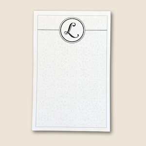 May include: White stationery notepad with a decorative floral pattern. The top features a black circular emblem with a stylized letter "L". The notepad has a thin black border.