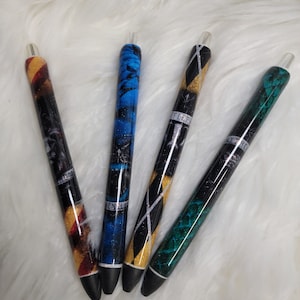 May include: Four pens with different designs. The pens are black with glitter and have different colored stripes. The designs include a green snake, a gold and black pattern, a blue and black pattern, and a red and gold pattern. The pens have clear tops and black tips.