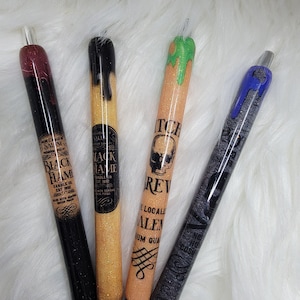 May include: Four decorative pens with glitter and themed designs. The pens feature the text "Black Flame Candle Co." and "Witch's Brew" with skull graphics. The pens have black, gold, orange, and blue accents, with a clear tip.