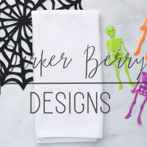 May include: A white kitchen towel with the text "Parker Berry Designs" written in black cursive script. Three plastic skeleton figurines in orange, green, and purple are arranged around the towel. A black paper spiderweb is in the upper left corner of the image.