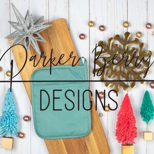 Puede incluir: Un agarrador de olla de color turquesa con el texto "Parker Berry Designs" en una tabla de cortar de madera con un copo de nieve dorado y pequeños árboles de Navidad.