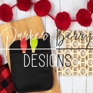 May include: Black oven mitt with two red and green kitchen utensils inside. The oven mitt is on a wooden cutting board with a gold "HO HO HO" decorative mat and red pom poms in the background. The text "Parker Berry Designs" is written on the oven mitt.