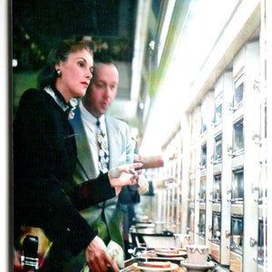 May include: A woman in a black suit jacket and a man in a patterned shirt stand in front of a vending machine. The woman is holding a tray with food on it. The vending machine has many rows of small compartments.