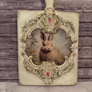 May include: A rectangular cream-colored decorative plaque featuring an ornate, raised relief frame with scrollwork and two small pink rose accents. The center displays a detailed illustration of a brown rabbit against a light, cracked background.