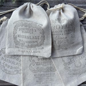 Puede incluir: Una colección de bolsas con cordón de color beige hechas de tela tejida. Cada bolsa presenta una etiqueta impresa de estilo vintage en tinta gris oscuro, que muestra varios textos, incluidos Josephine's Famous Marmalade y Royal Lemon Curd.