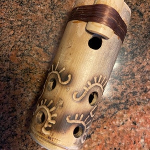 May include: A brown bamboo tube with a carved design of eyes and a brown leather strap. The tube has several holes drilled into it.