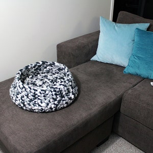 May include: A black, white, and gray knitted pet bed sits on a gray couch. The couch has three pillows, one light blue, one teal, and one white with a squirrel design.