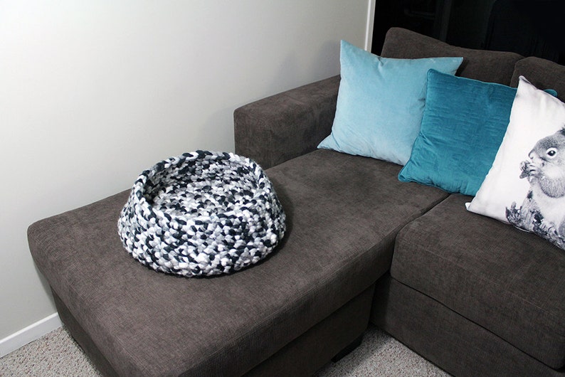 May include: A black, white, and gray knitted pet bed sits on a gray couch. The couch has three pillows, one light blue, one teal, and one white with a squirrel design.