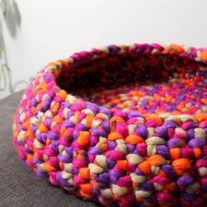May include: A colorful crocheted pet bed made with pink, orange, purple, and beige yarn. The bed is round and has a deep well for a pet to rest in.
