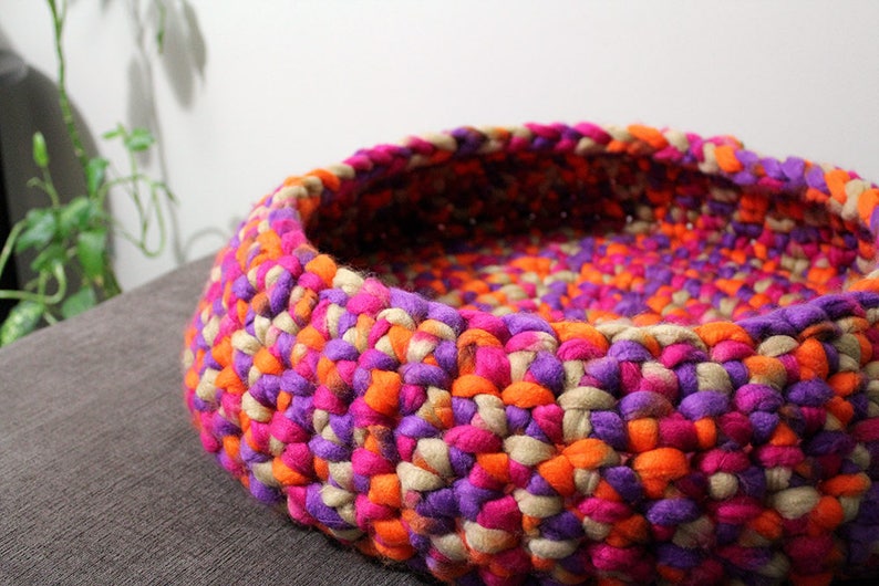 May include: A colorful crocheted pet bed made with pink, orange, purple, and beige yarn. The bed is round and has a deep well for a pet to rest in.
