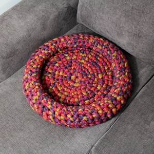 May include: A round, crocheted pet bed in shades of red, purple, and yellow sits on a gray couch.
