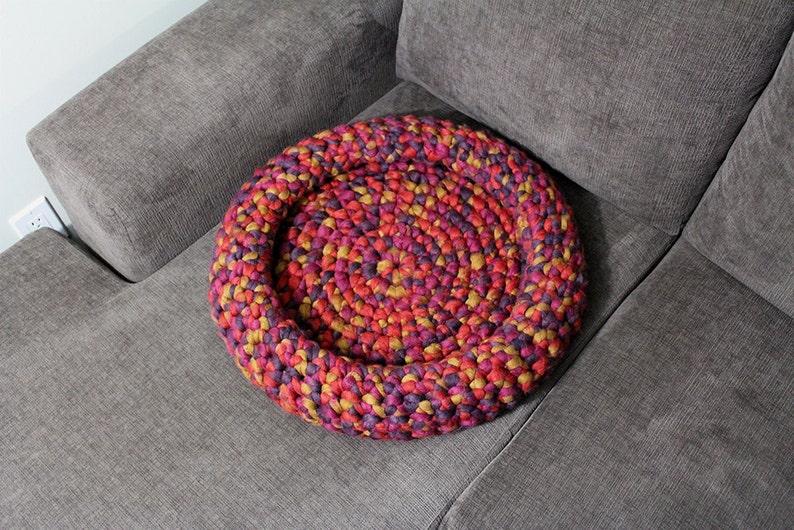 May include: A round, crocheted pet bed in shades of red, purple, and yellow sits on a gray couch.