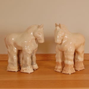 Puede incluir: Dos esculturas de caballos de cerámica de color crema. Los caballos se enfrentan, con detalles precisos y un acabado brillante. Se colocan sobre una superficie de madera. Cada escultura mide unos 30 cm de altura.