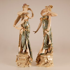 May include: Two porcelain figurines of women in flowing gowns with gold accents. The women are standing on rocky bases and are holding their gowns with their hands.