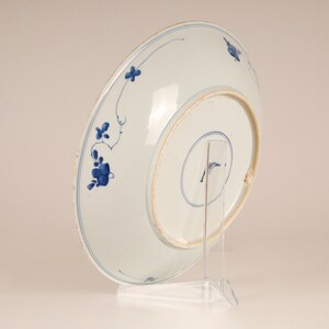 May include: A white porcelain plate with a blue and white floral design. The plate is on a clear stand.