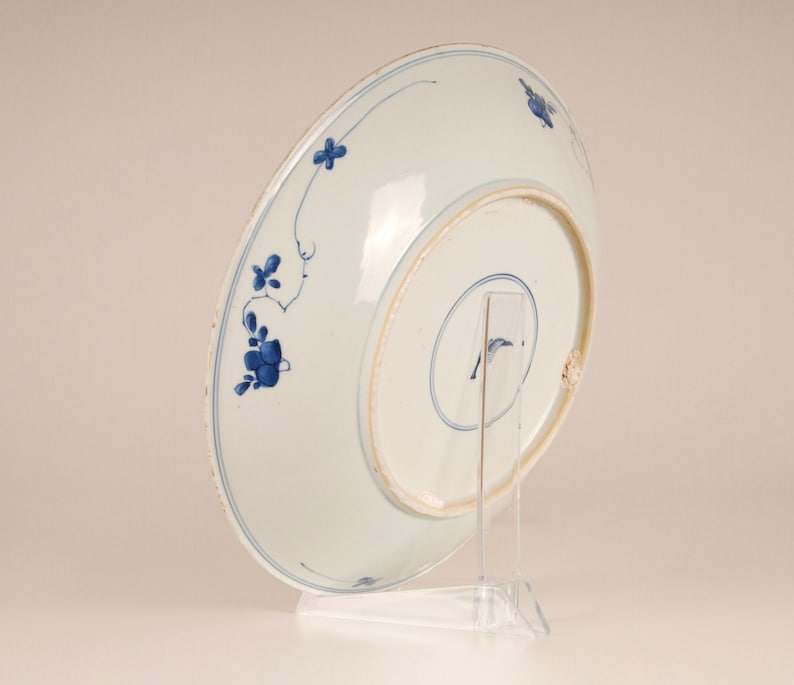 May include: A white porcelain plate with a blue and white floral design. The plate is on a clear stand.