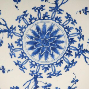 May include: A white ceramic plate with a blue and white floral design. The centre of the plate features a large blue flower with a white centre. The border of the plate is decorated with a repeating pattern of blue flowers and leaves.