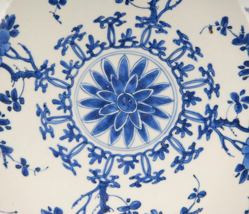 May include: A white ceramic plate with a blue and white floral design. The centre of the plate features a large blue flower with a white centre. The border of the plate is decorated with a repeating pattern of blue flowers and leaves.