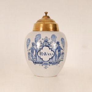 Może przedstawiać: Biały ceramiczny dzbanek z niebiesko-białym wzorem przedstawiającym postacie i napis "HAVANA". Dzbanek ma mosiężną pokrywkę.