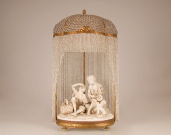 Puede incluir: Una base de lámpara de porcelana blanca con dos figuras de niños sentados bajo un dosel de cuentas de cristal. La base es de color dorado y tiene forma redonda. La lámpara está sobre un fondo blanco.