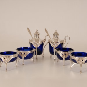 May include: An antique silver and cobalt blue glass serving set. The set comprises two lidded containers with spoons and four open bowls. The silver rims and legs feature intricate detailing. The cobalt blue glass provides a striking contrast.