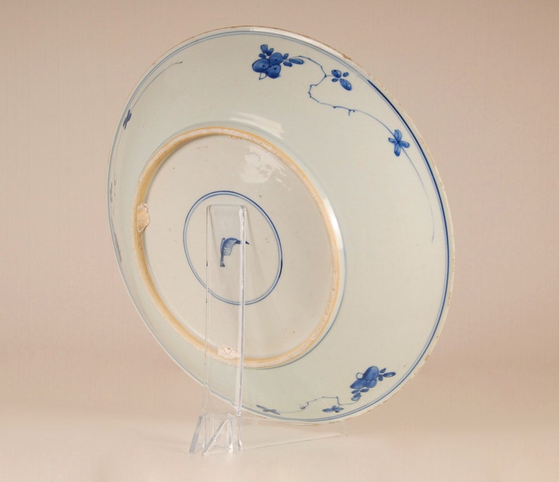 May include: A white porcelain plate with a blue and white floral design. The plate is on a clear plastic stand.