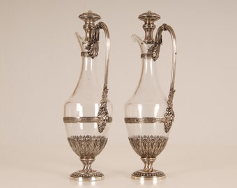 May include: A pair of silver and glass decanters with ornate details. The decanters have a clear glass body with a silver base, handle, and spout. The silver is decorated with a floral pattern.