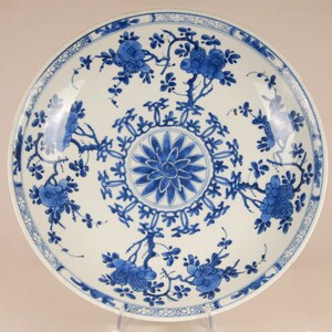 May include: A large, white porcelain plate with a blue and white floral design. The plate features a central medallion with a stylised flower and a repeating border of geometric shapes. The rim of the plate is decorated with a continuous floral motif.
