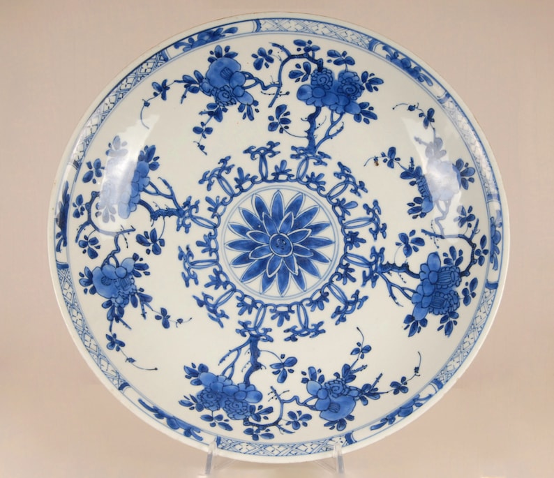 May include: A large, white porcelain plate with a blue and white floral design. The plate features a central medallion with a stylised flower and a repeating border of geometric shapes. The rim of the plate is decorated with a continuous floral motif.