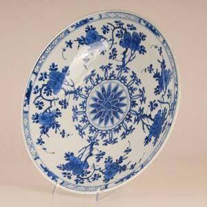 May include: A large, white porcelain plate with a blue and white floral design. The plate has a central medallion with a stylised flower and a border of repeating floral motifs.