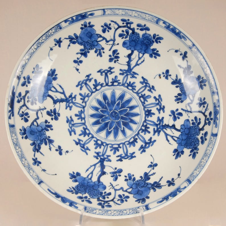 May include: A large, white porcelain plate with a blue and white floral design. The plate has a central medallion with a stylised flower and a border of geometric patterns. The floral design features branches with leaves and flowers.