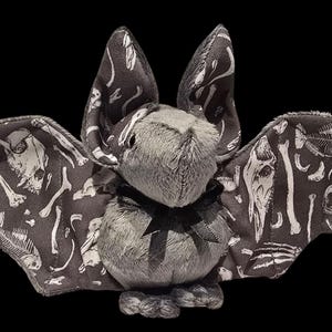 May include: A plush bat toy with outstretched wings. The bat's body is gray, with a black bow. The wings are black with a white bone pattern. The bat has large ears and a friendly expression.