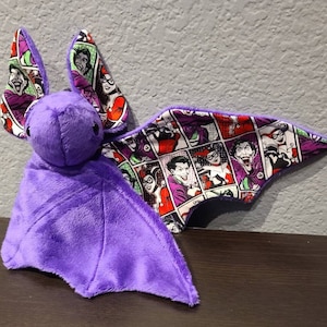 May include: A plush purple bat with comic book-themed wings and ears. The wings and ears feature a collage of comic book panels with characters in purple, red, and white. The bat's body is a solid, soft purple.